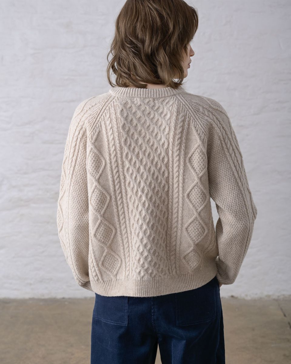 Back view of a woman wearing the BIBICO Derby Aran Cardigan in Oat, showing the full all-over Aran cable knit in diamond and rope cable panels, raglan sleeve seams, ribbed hem, and relaxed silhouette in warm oat lambswool, styled with navy wide-leg pants against a whitewashed brick wall