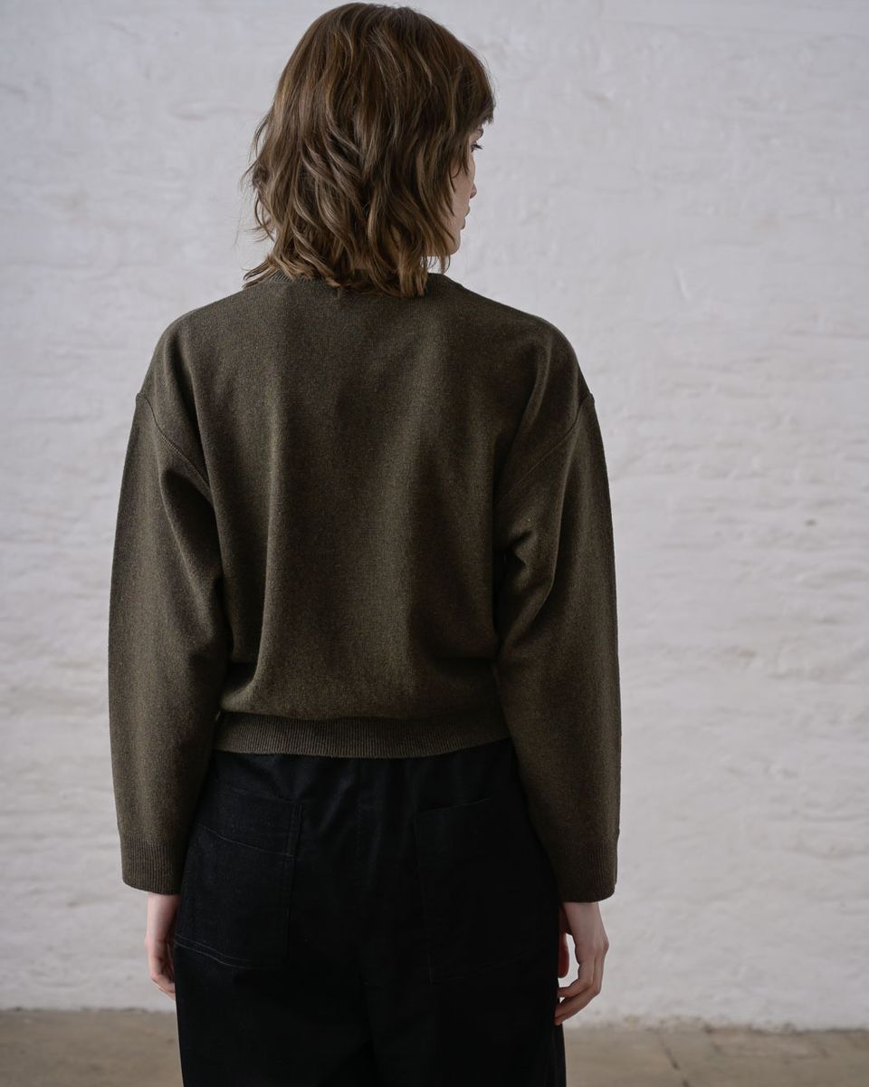 Back view of a woman wearing the BIBICO Ona Jumper in Olive, showing the dropped shoulders, wide long sleeves, wide ribbed hem band, and cropped silhouette in lightweight olive Geelong Merino wool, styled with black wide-leg trousers against a whitewashed brick wall