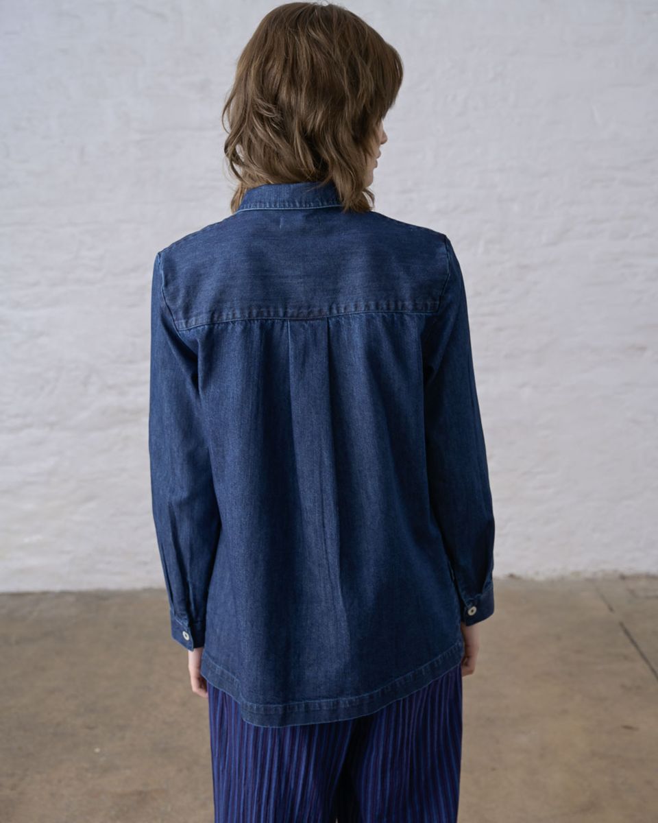 Back view of a woman wearing the BIBICO Miriam Tunic in Denim, showing the back yoke, soft box pleats below the yoke, full-length sleeves with buttoned cuffs, curved shirttail hem, and relaxed silhouette in deep indigo 9oz cotton denim, styled with a blue pleated skirt against a whitewashed brick wall