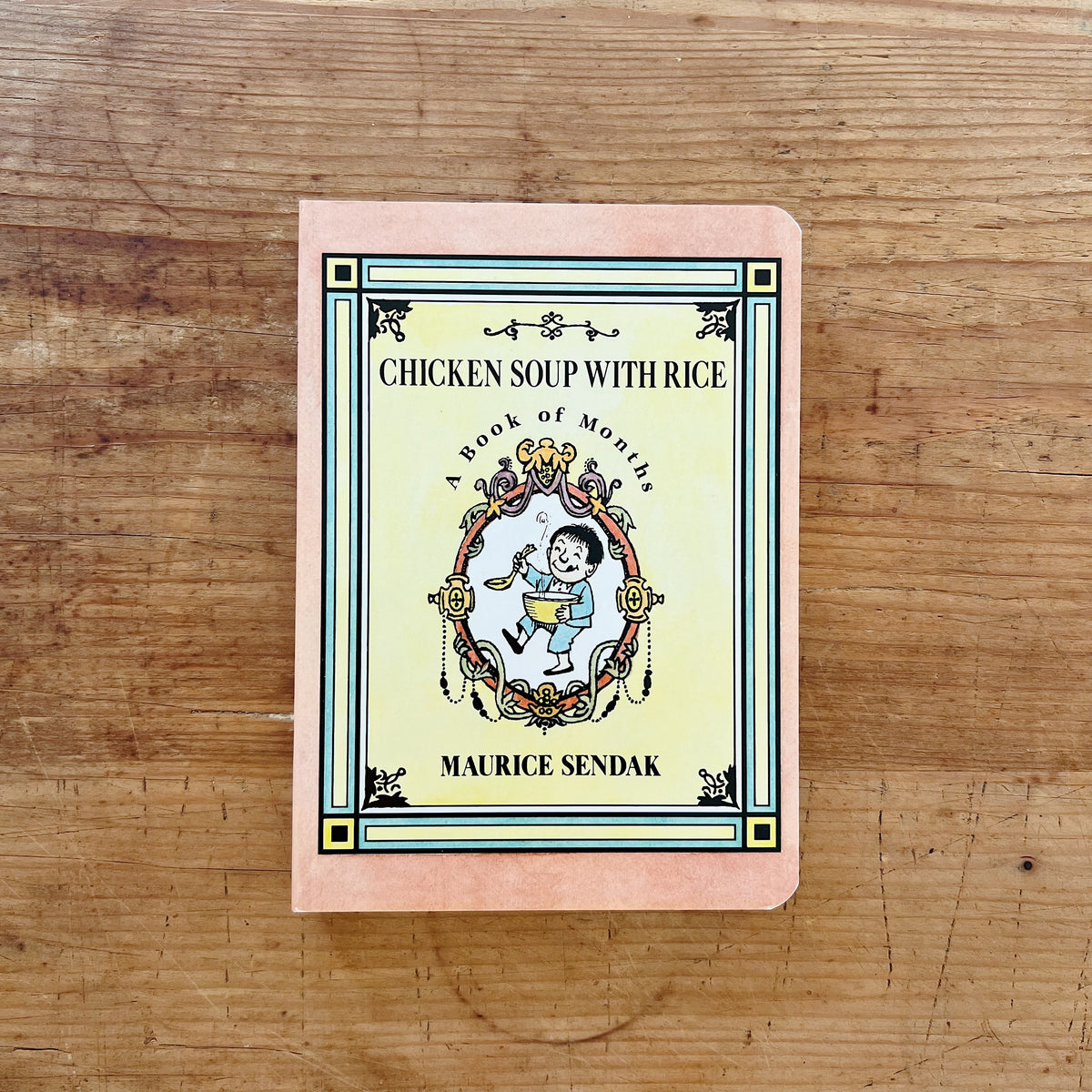 Front cover of Chicken Soup with Rice: A Book of Months by Maurice Sendak, featuring a pink cover with a decorative ornamental border framing a boy joyfully eating soup from a bowl, with the title in bold lettering and the author's name at the bottom, displayed flat on a warm wood surface.