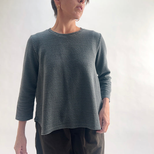 Close front view of model wearing an iron charcoal and black tonal textured stripe crewneck top with 3/4 sleeves, arm at side, showing the crew neckline, crinkle-texture fabric, and relaxed A-line silhouette, styled with dark olive wide-leg pants.