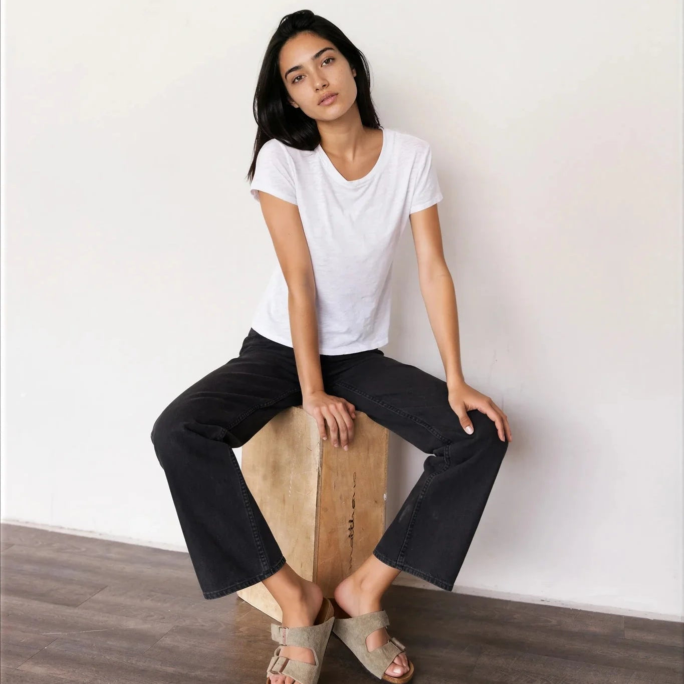 Model wearing It Is Well Cropped Tee in Salt, seated on a wooden block against a white wall, front view showing crisp white lightweight slub jersey, clean crew neck, short fitted sleeves with slight cuff, and slightly cropped length in slim petite fit, styled with black wide-leg cropped pants and taupe Birkenstock slide sandals