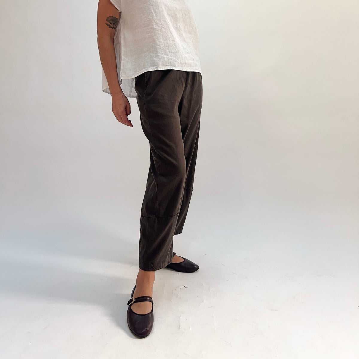 Side view of walnut linen-tencel lantern pants showing tapered curved leg, ankle-length hem seam detail, and elastic waistband, paired with a white linen high-low top and dark Mary Jane flats