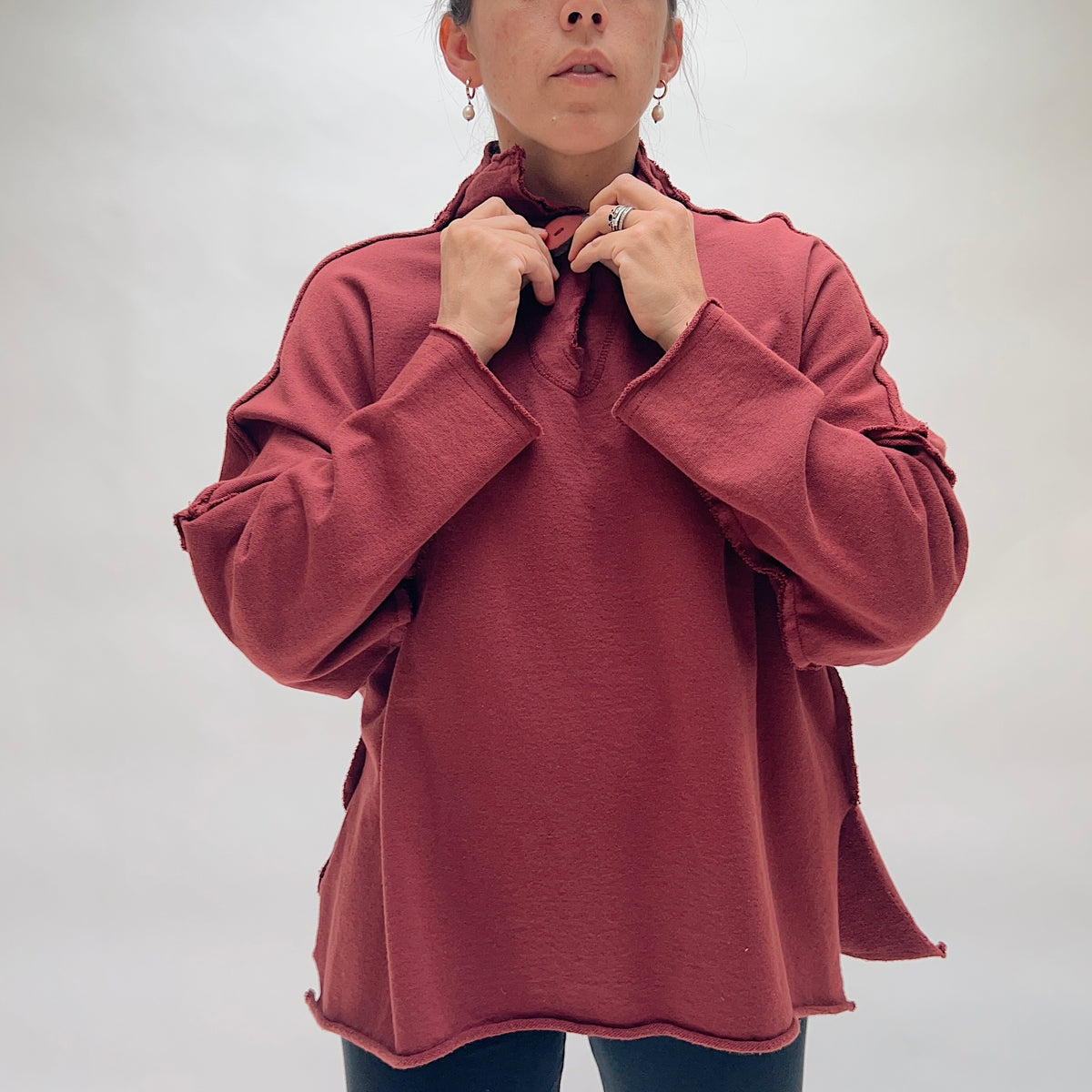 Close front view of the Eleven Stitch Long Rib Out Funnel Neck Slub Sweatshirt in Berry, with model's hands pulling up the ruffled funnel neck, highlighting the single large button closure, exposed raw rib seams at the shoulders and cuffs, and the raw-edge curved hem of the oversized berry slub cotton sweatshirt