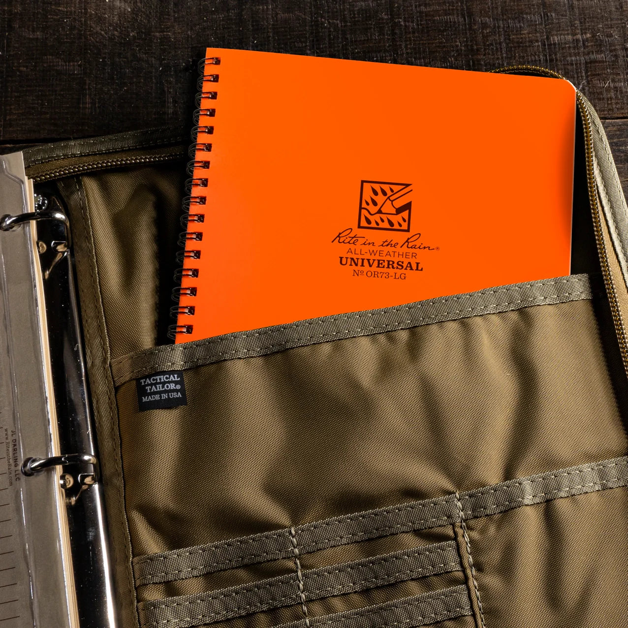 An orange spiral-bound notebook with black printed text and logo, partially tucked into a zippered pocket of an olive green binder. The notebook cover shows the text 'Kit in My Heart UNIVERSAL NY 9876-LOE' and a small square logo.