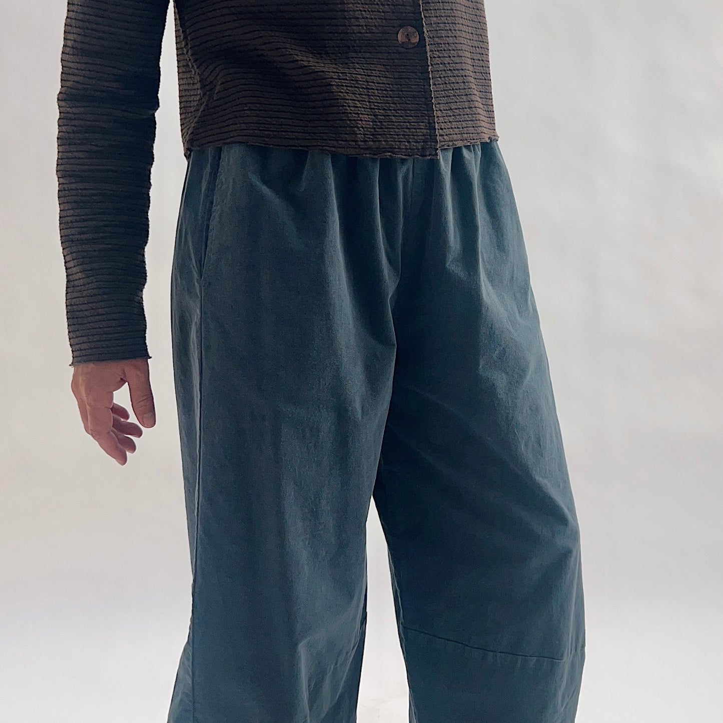 Close front view of model wearing iron blue-gray mini cord lantern pants, showing the gathered elastic waist, fine cord texture, and wide lantern volume at the hip, styled with the matching chocolate striped cropped cardigan against a white background.