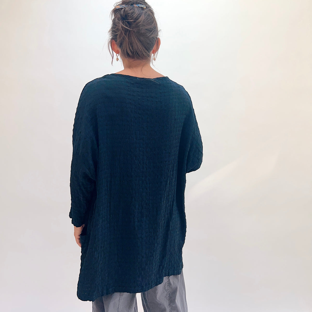 Back view of teal crinkle-texture one-size pocket tunic showing relaxed boxy silhouette, scoop back neckline, and 3/4-length sleeves, hair up with clip, styled with grey wide-leg pants
