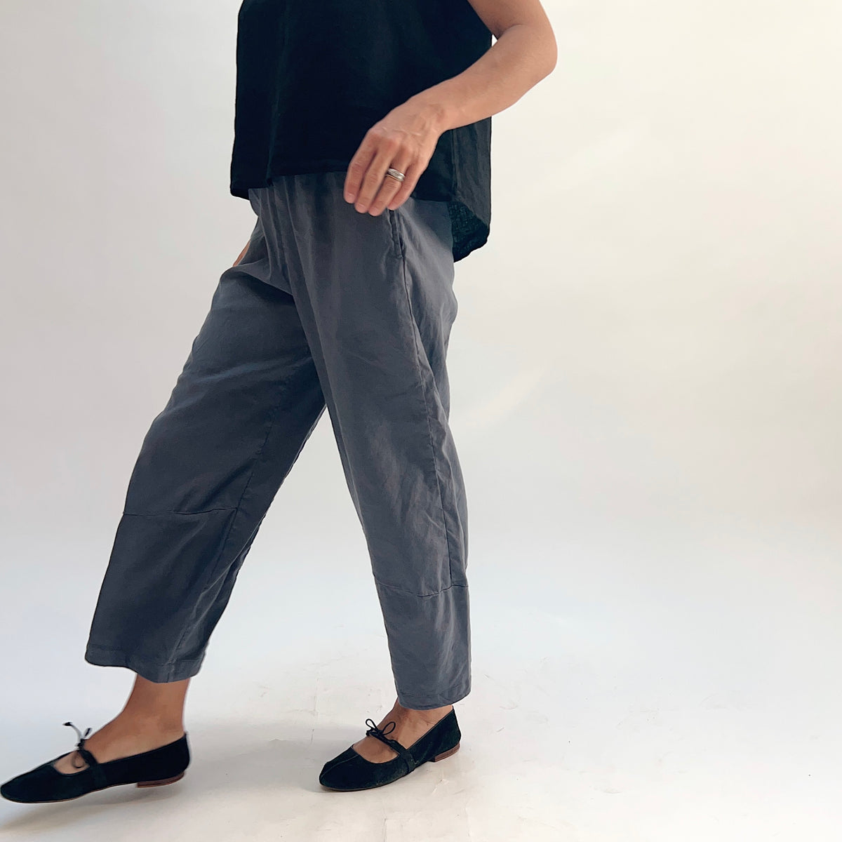Side view of pewter linen-tencel lantern pants showing curved seam leg, side pocket, and elastic waistband, worn with a black linen top and black bow-tie ballet flats, hand resting near pocket
