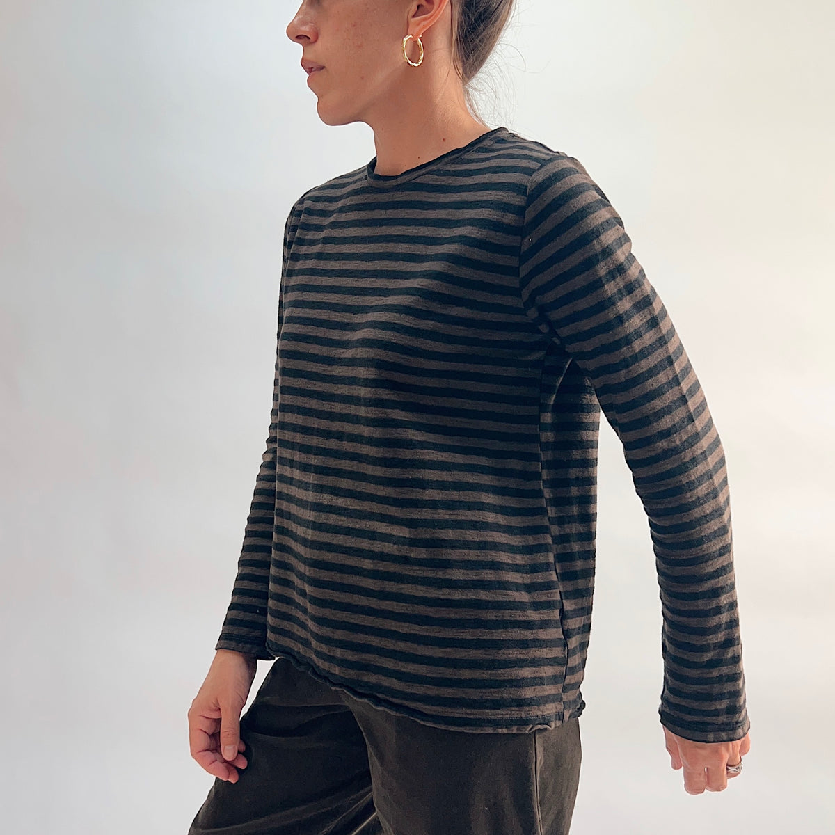 Side profile of model wearing the Cut Loose Striped Long Sleeve Easy Tee in Chocolate, showing the crew neckline, relaxed easy silhouette, long sleeves, and curved hem in a lightweight chocolate and black horizontal stripe cotton-linen blend, styled with the chocolate wide-leg crop trouser