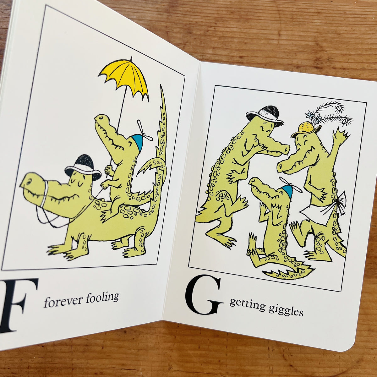 Open interior spread of Alligators All Around showing two pages: on the left, the letter F with a caption "forever fooling" and an illustration of two alligators in bowler hats — one riding the other's back while holding a yellow umbrella; on the right, the letter G with a caption "getting giggles" and an illustration of three alligators in hats laughing and dancing together, displayed on a warm wood surface.