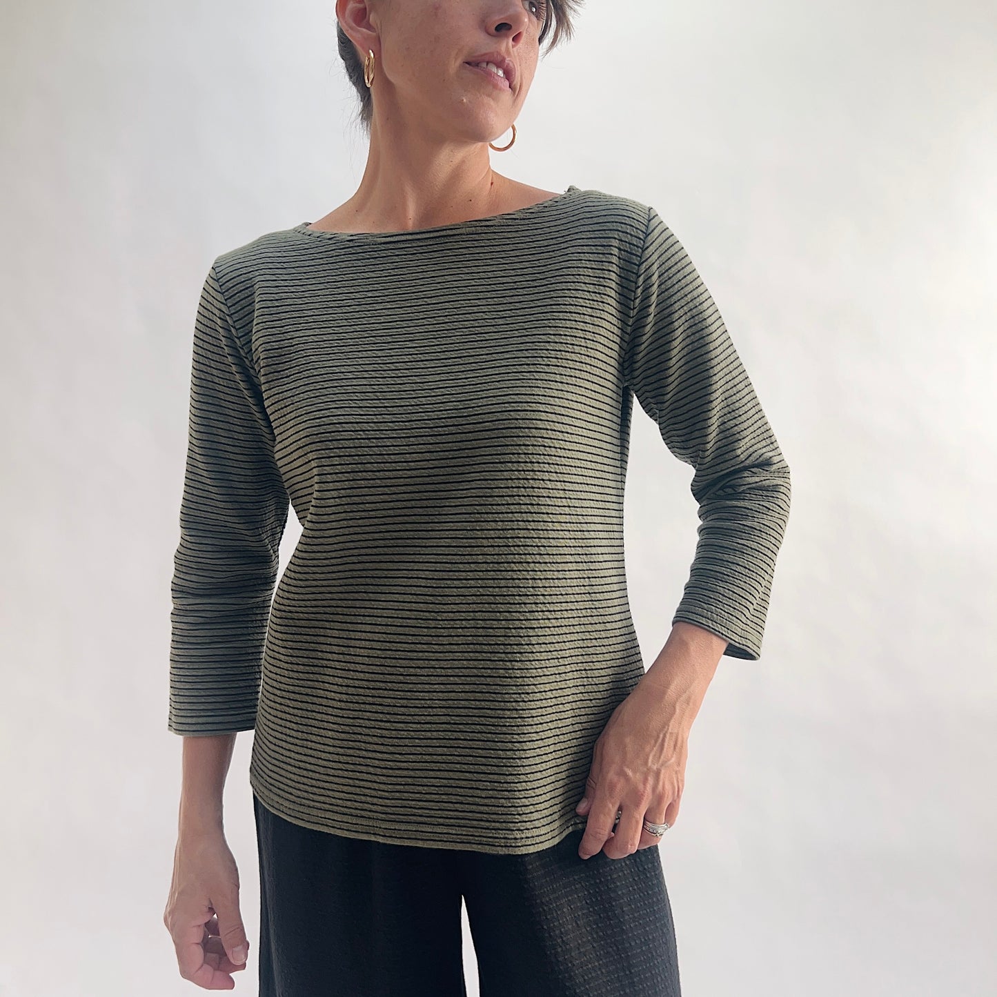 Full front view of model wearing a kelp green tonal textured stripe boatneck tee with 3/4 sleeves, hand on hip, showing the relaxed fit, wide boatneck, and crinkle-texture fabric, styled with black wide-leg pants against a white background.