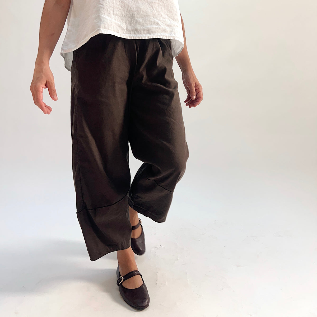 Close-up front view of walnut linen-tencel lantern pants showing wide curved leg silhouette, elastic waistband, and cropped ankle length with curved hem seam detail, styled with a white linen top and dark Mary Jane flats