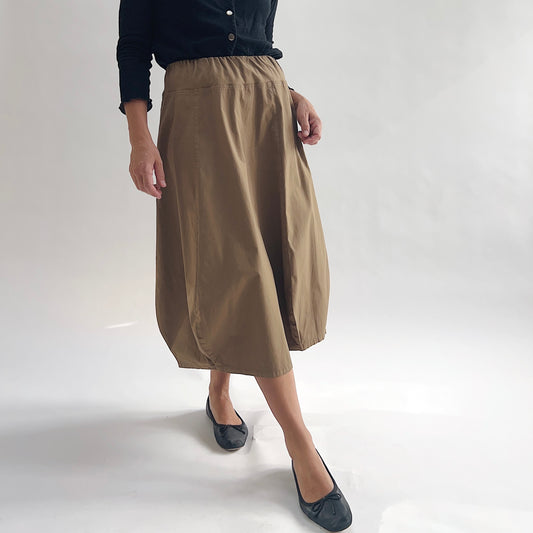 Three-quarter side view of model wearing Eleven Stitch winter olive rib yoke skirt in motion, showing elastic knit waistband, curved seam paneling, sculptural bubble hem, below-the-knee length, and warm khaki olive color, styled with black long sleeve top and black ballet flats