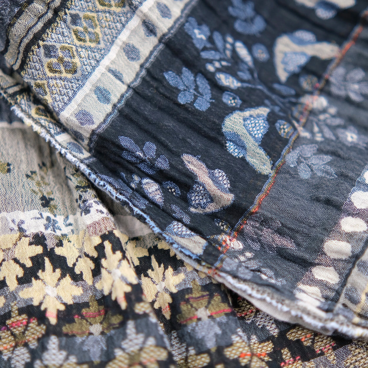 Close-up detail of the Létol Anoushka Scarf in Noir, showing the richly textured Jacquard weave where both sides of the scarf are visible in layered folds — the black ground side with gold and oatmeal oak leaf and floral motifs alongside the oatmeal ground side with chambray blue botanical and paisley patterns, with a red accent selvedge edge visible between the layers, highlighting the dimensional woven construction and tonal contrast between the two sides
