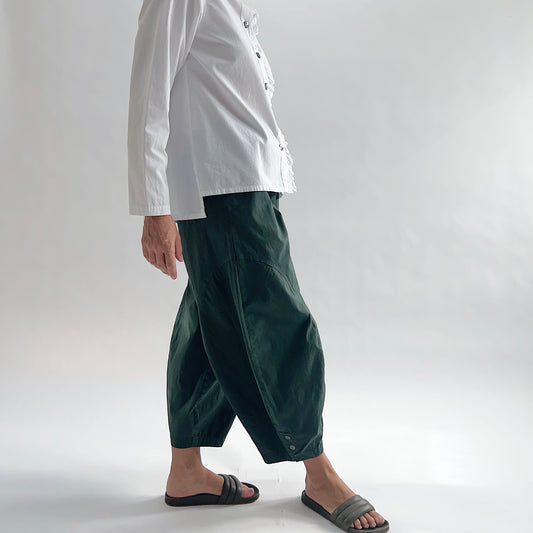 Side view of model wearing Eleven Stitch konbu snap doreen pant in motion, showing elastic waistband, wide balloon leg tapering to snap hem at ankle, side seam pocket, and deep forest green color, styled with white blossom collar shirt and dark slide sandals