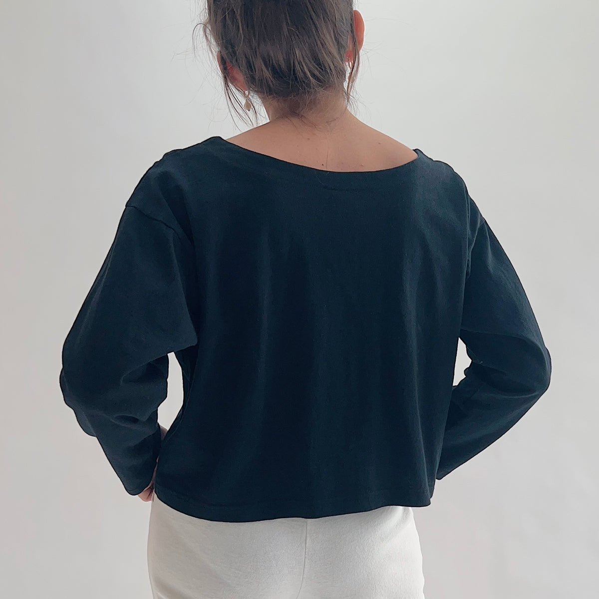 Back view of model wearing Pacific Cotton black long sleeve button front shirt, showing wide scoop back neckline, long sleeves, boxy cropped silhouette, banded hem, and black color