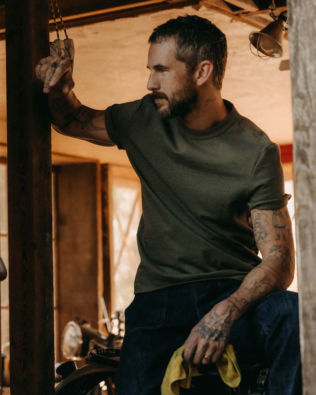 Man wearing the Taylor Stitch Organic Cotton Shop Tee in Army Twist, three-quarter lifestyle shot in a rustic workshop, reaching overhead, styled with dark denim jeans