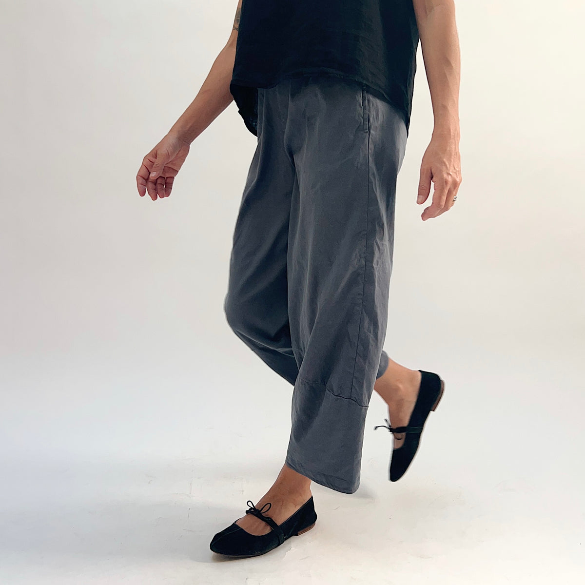 Dynamic front view of pewter linen-tencel lantern pants with wide barrel-leg silhouette, curved seam detail, and ankle-length hem, styled with a black linen top and black bow-tie ballet flats, one leg raised