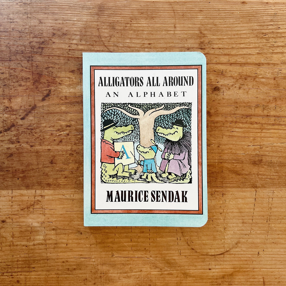 Front cover of Alligators All Around: An Alphabet board book by Maurice Sendak, featuring a mint green cover with a terracotta-bordered illustration of three cartoon alligators — one in a red jacket holding a card with the letter A, one small alligator in blue, and one in a purple robe — surrounded by a sketchy tree-like background, with the title in bold black lettering and the author's name at the bottom, displayed flat on a warm wood surface.