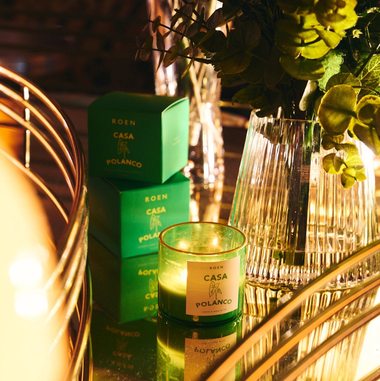 Roen Casa Polanco candle glowing in a green glass jar beside its green gift box, reflected on a mirrored tray with crystal glassware and warm ambient lighting