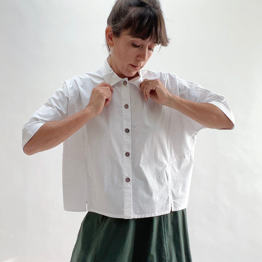 Full front view of model wearing Eleven Stitch chalk camp shirt with hands at collar, showing rounded collar, dark shell buttons, short sleeves, slightly cropped boxy fit, and white color, styled with forest green wide-leg skirt