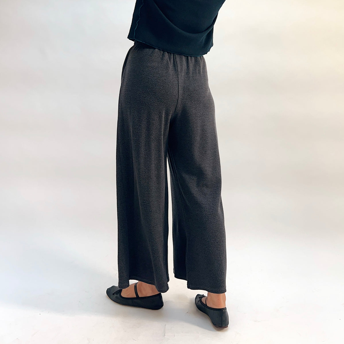 Model wearing the Cut Loose Marled Knit Crop Pant in Iron, back three-quarter view, showing elastic waistband, wide-leg silhouette, clean cropped hem, and subtle marled texture of dark charcoal knit, styled with a dark top and black mary jane flats