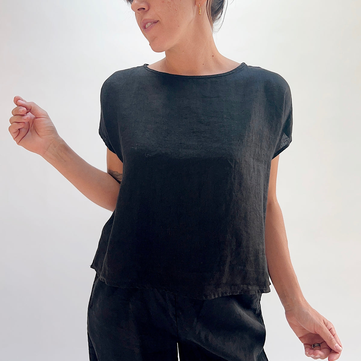 Front three-quarter view of black linen high-low tee clearly showing high-low hem, boatneck neckline, and cap sleeves, styled with black linen lantern pants