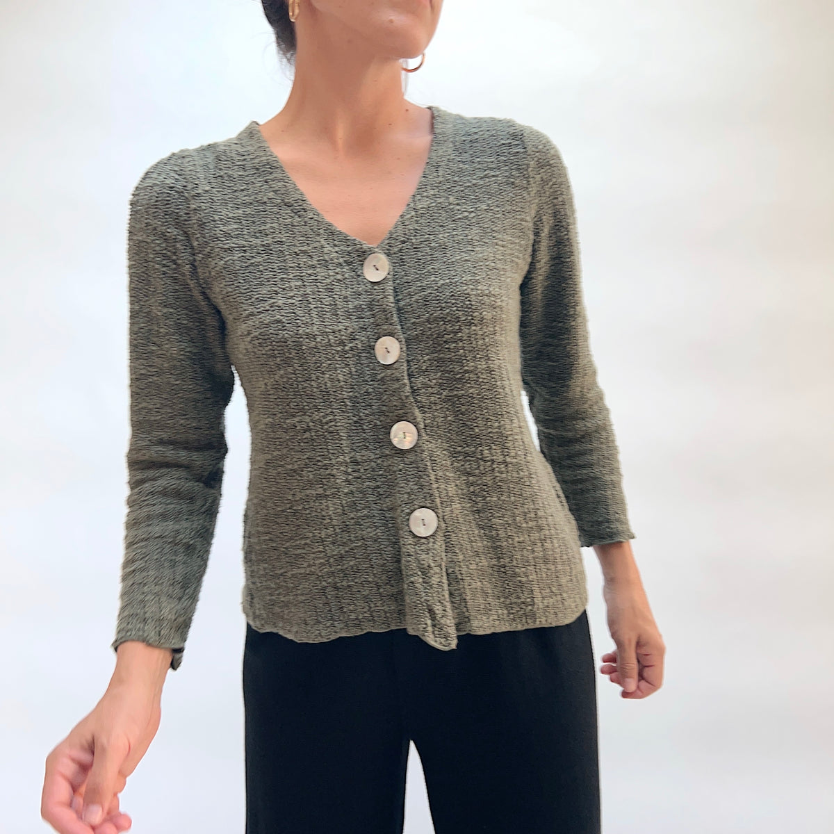 Model wearing the Cut Loose Puff Sleeve Cardigan in Kelp, three-quarter front view, olive green marled knit cardigan with V-neckline, four large natural shell buttons, gently puffed shoulders, and scalloped lettuce hem, styled with black pants