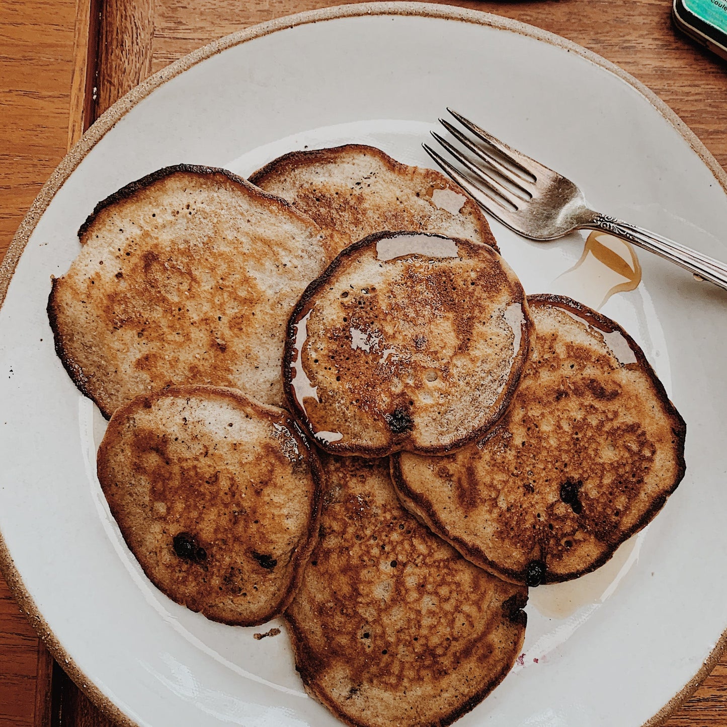 Six golden-brown pancakes with toasted edges arranged on a white ceramic plate with a beige rim, topped with a drizzle of syrup. A silver fork rests on the upper right side of the plate, which sits on a wooden table.
