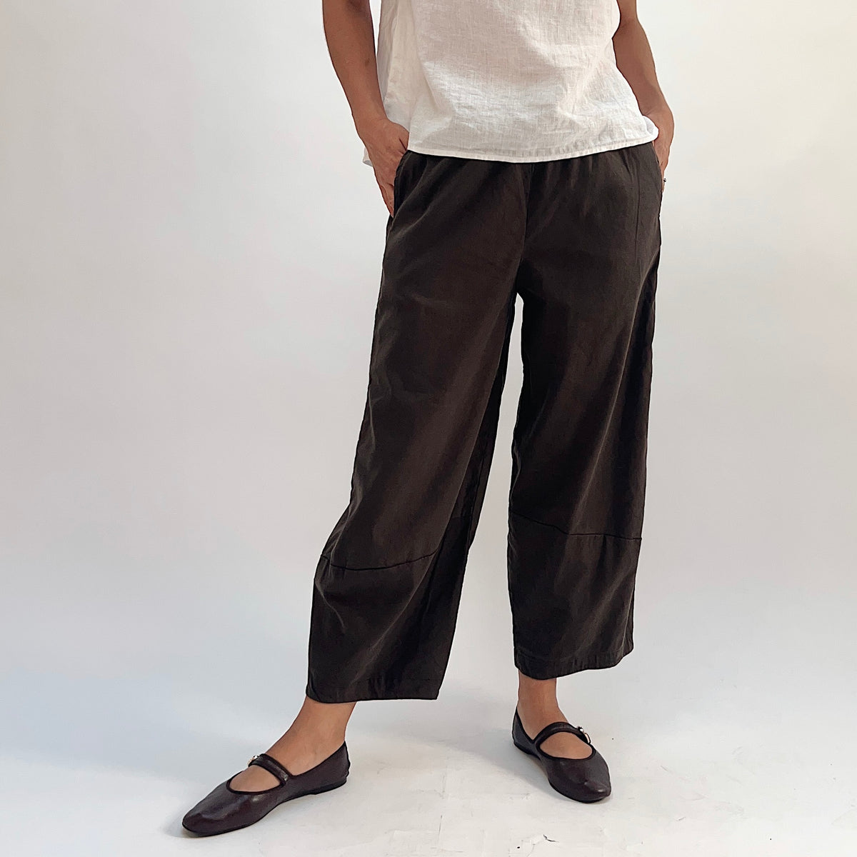 Full front view of walnut linen-tencel lantern pants with wide curved leg, elastic waistband, side pockets, and curved ankle hem seam, styled with a white linen top and dark Mary Jane flats, hand in pocket