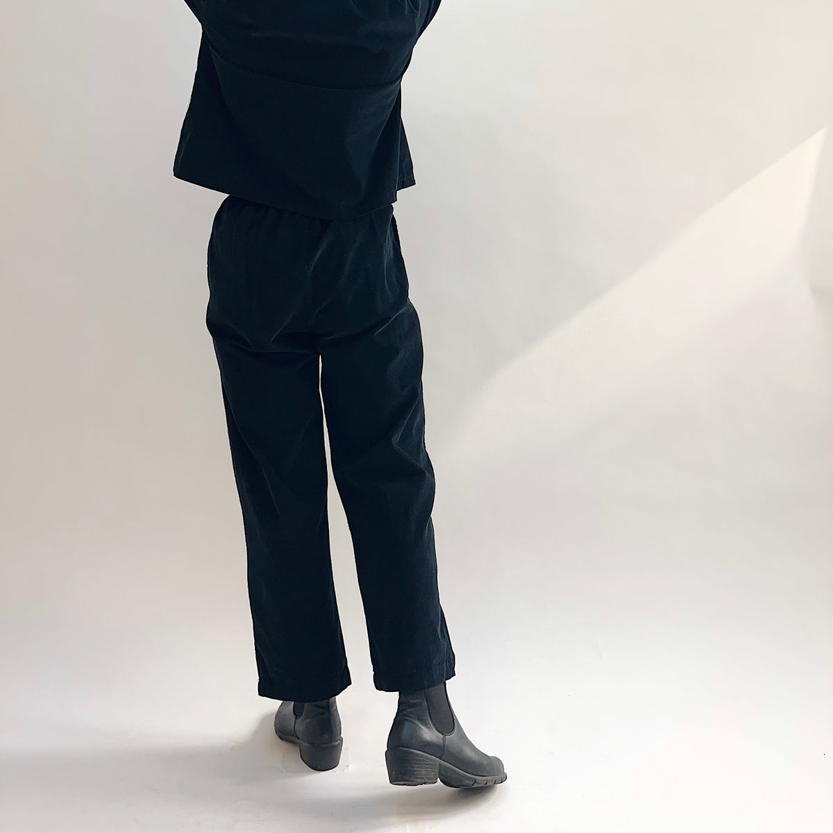 Back view of model wearing the Bryn Walker Corduroy Pleated Pants in Black, showing elastic back waistband, straight leg, full-length hem, and black pinwheel corduroy drape, styled with matching jacket and black Chelsea boots