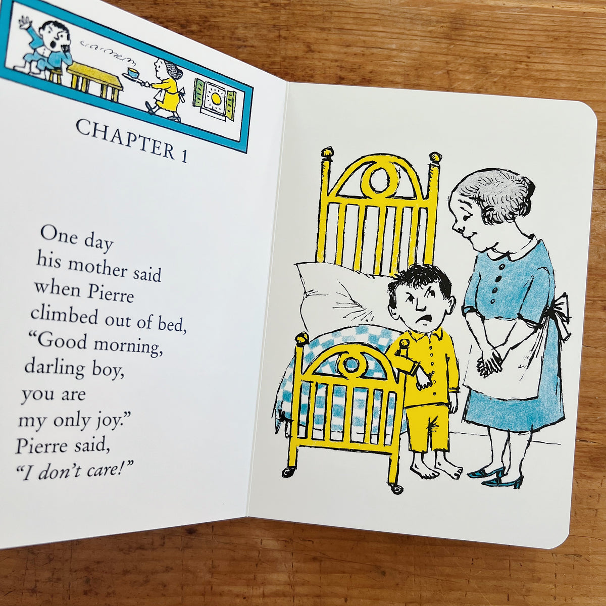 Open interior spread of Pierre board book displayed on a warm wood surface, showing Chapter 1. The left page has a small turquoise-bordered header illustration of a scene from the story, the chapter heading "CHAPTER 1" in bold black caps, and the verse text: "One day / his mother said / when Pierre / climbed out of bed, / 'Good morning, / darling boy, / you are / my only joy.' / Pierre said, / 'I don't care!'" The right page shows Maurice Sendak's full-color illustration of Pierre — a small boy in yellow pa