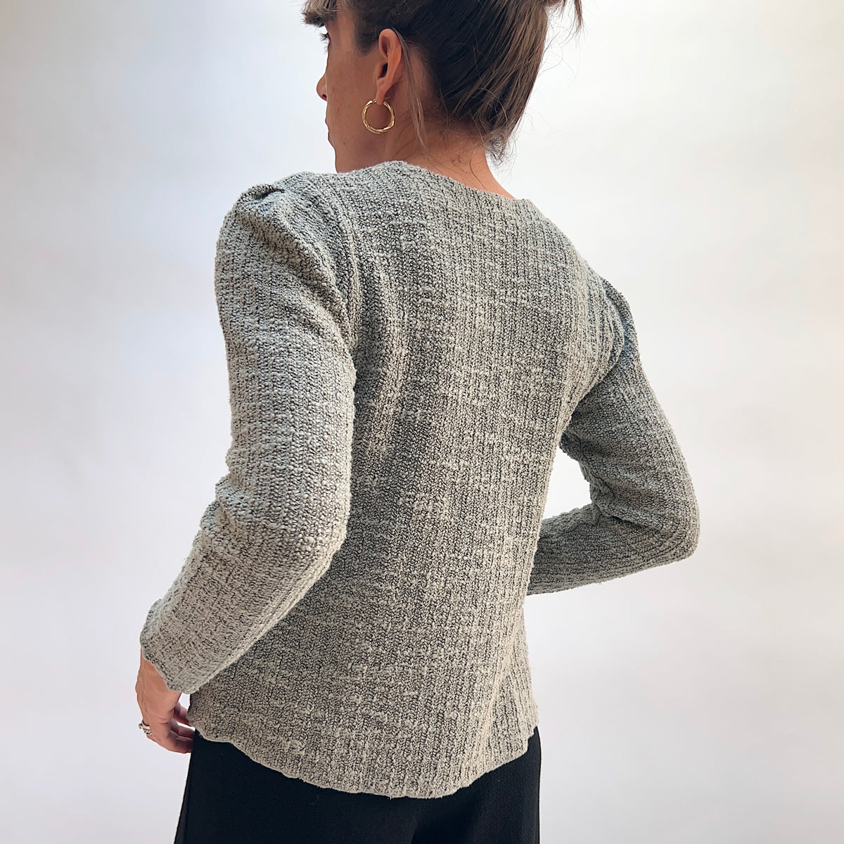 Model wearing the Cut Loose Puff Sleeve Cardigan in Squirrel, back view with hands behind back, showing gently puffed shoulder seam, ribbed back panel, and scalloped lettuce hem of warm grey-taupe marled knit cardigan, styled with black pants
