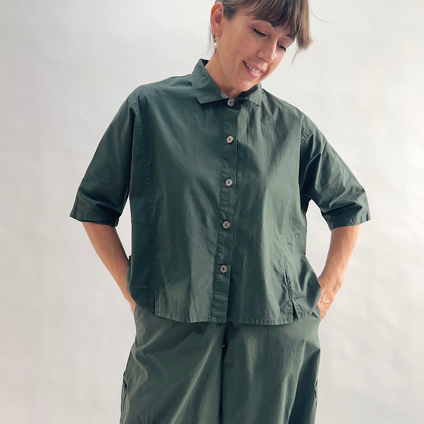 Full front view of smiling model wearing Eleven Stitch konbu camp shirt with hands in pockets, showing rounded collar, dark shell buttons, short sleeves with rolled cuffs, slightly cropped boxy fit, side slits, and deep forest green color, styled with matching konbu snap front pant