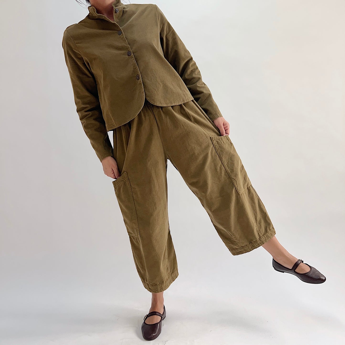 Model playfully posing in the Bryn Walker Corduroy Pasha Pants in Partridge, full-length front view showing elastic waistband, two side leg patch pockets, cropped wide-leg hem, and olive pinwheel corduroy, styled as a complete set with matching corduroy jacket and dark Mary Jane flats