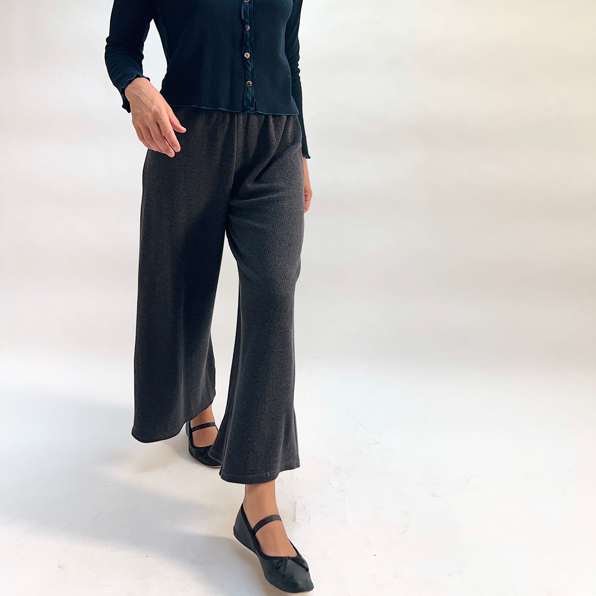 Model wearing the Cut Loose Marled Knit Crop Pant in Iron, three-quarter front view with hand in pocket, showing relaxed wide-leg fit, elastic waistband, and fluid drape of dark charcoal marled knit, styled with a dark teal cardigan and black mary jane flats