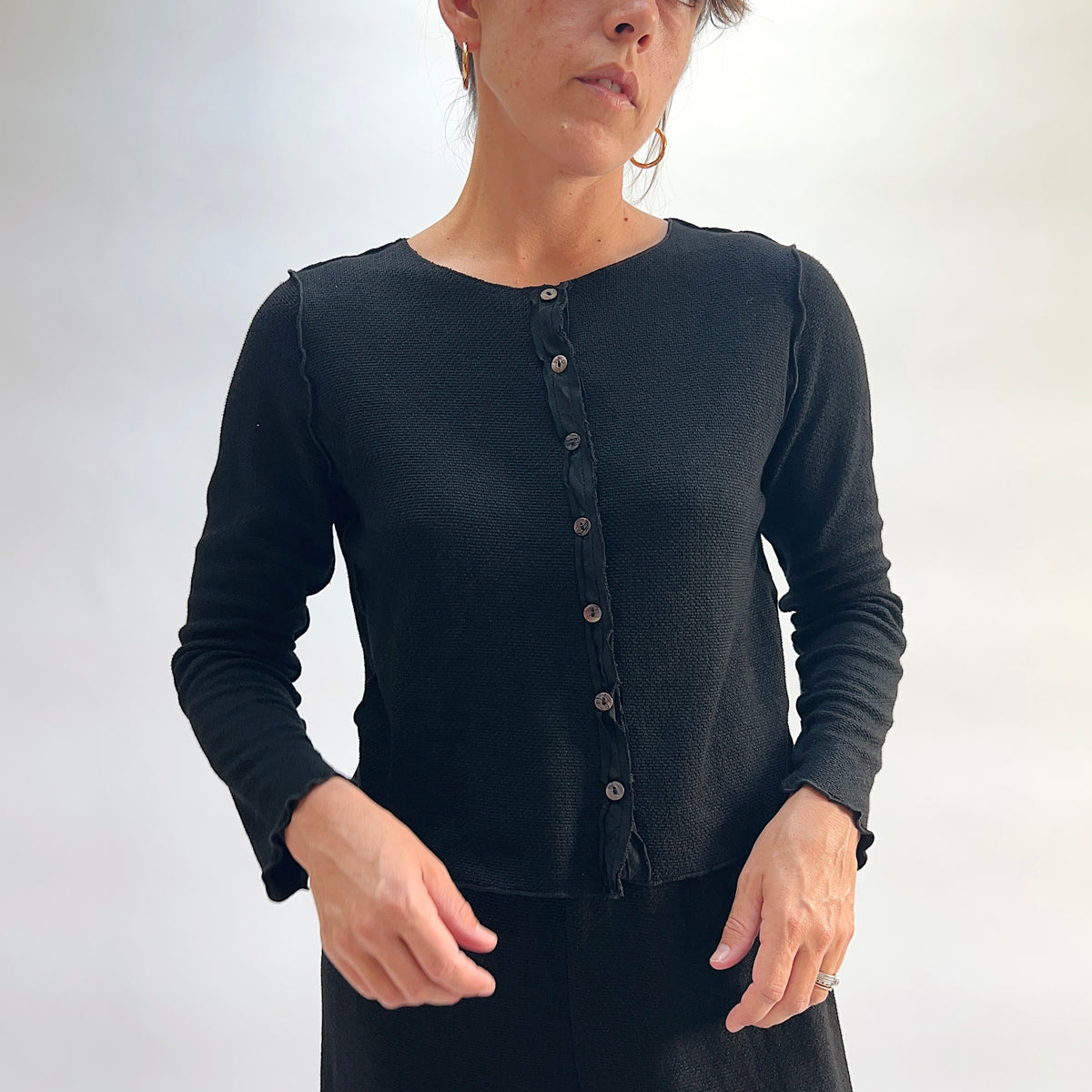 Model wearing the Cut Loose Marled Knit Cropped Cardigan in Black, full-length front view looking down, showing round neckline, scalloped lettuce edges, dark shell button placket, and relaxed cropped fit of black marled knit cardigan, styled with a black skirt