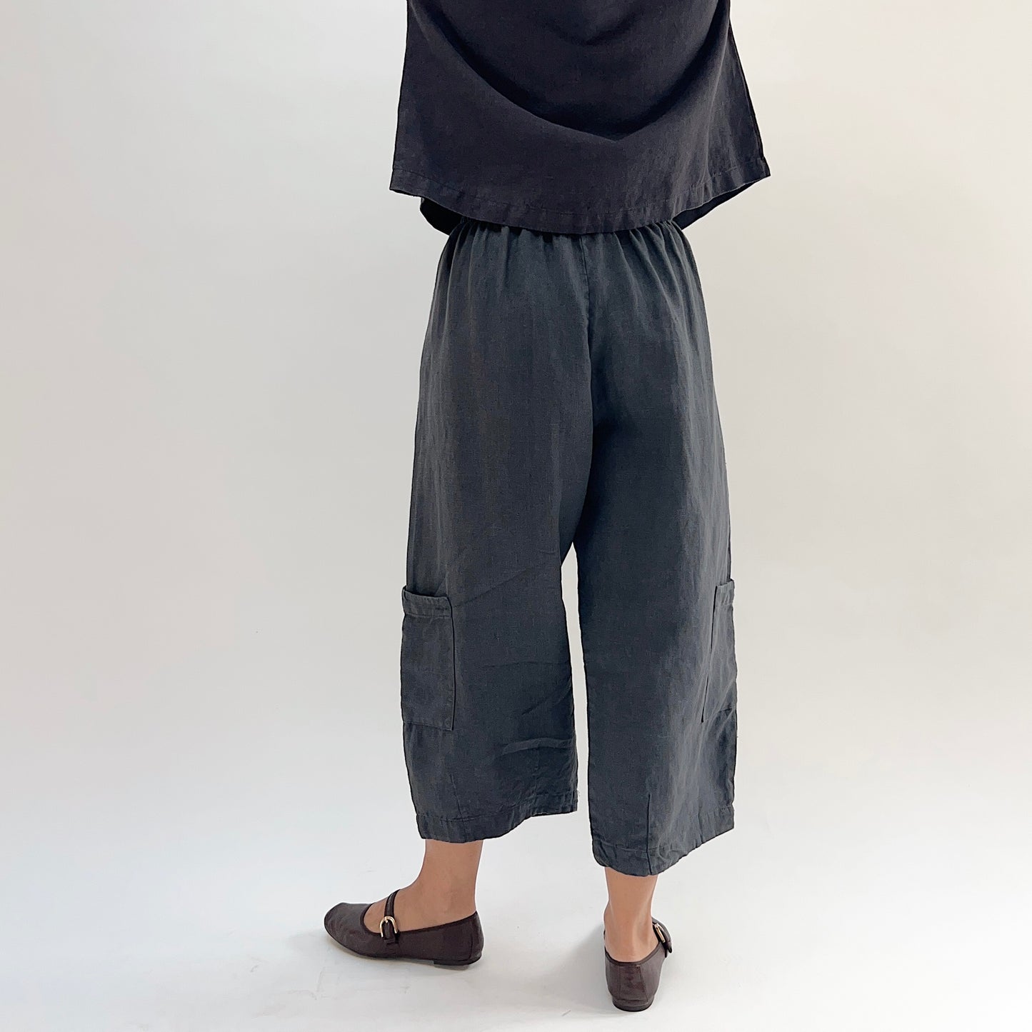 Back view of model wearing the Bryn Walker Heavy Linen Casbah Pant in Moonlight, showing fully elastic waistband, barrel-tapered leg, two large lower-leg patch pockets, cropped ankle length, and deep moonlight heavyweight linen, styled with matching linen top and brown Mary Jane flats