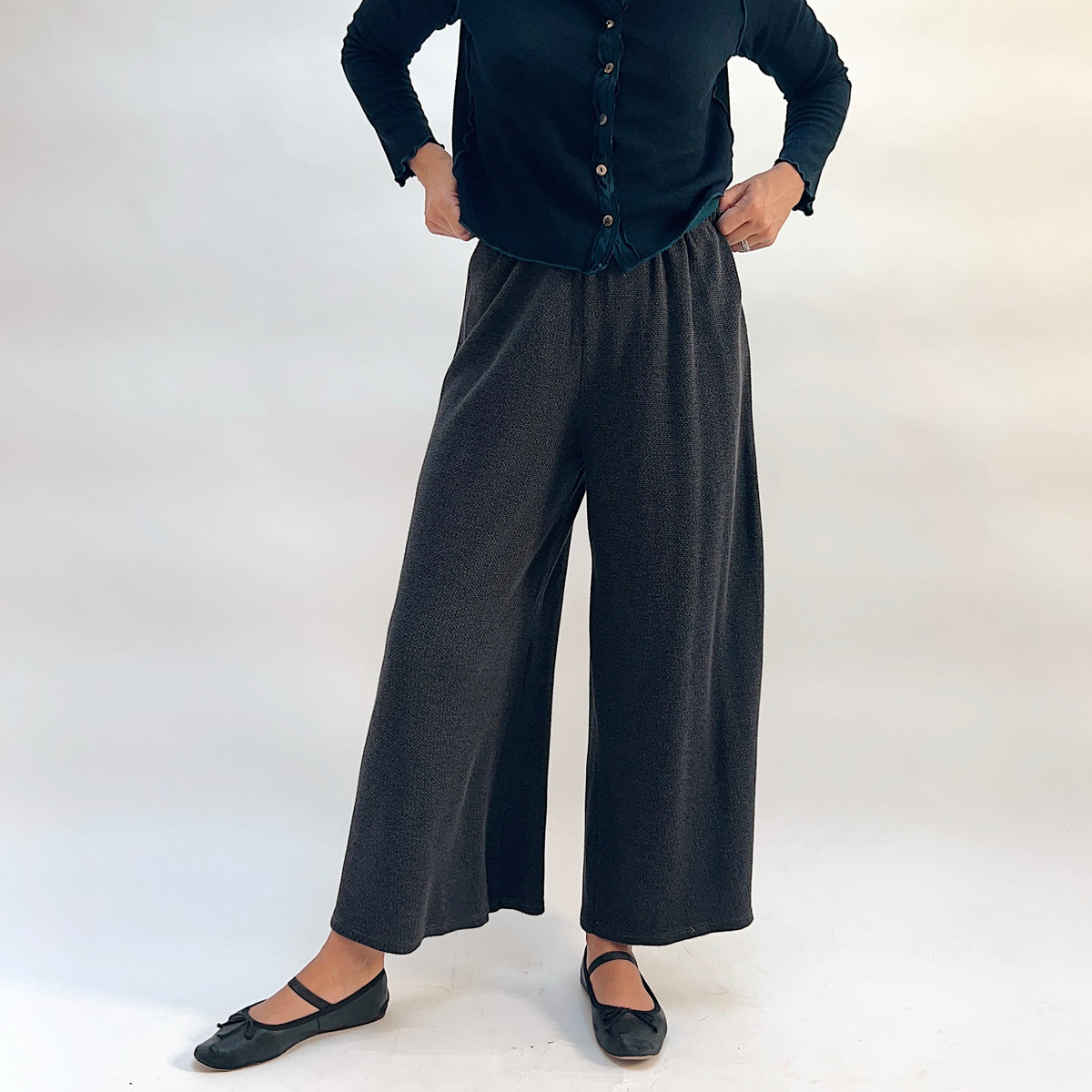 Model wearing the Cut Loose Marled Knit Crop Pant in Iron, full-length front view with hands on waist, showing wide-leg crop silhouette, elastic waistband, and dark charcoal marled knit, styled with a dark teal cardigan and black mary jane flats