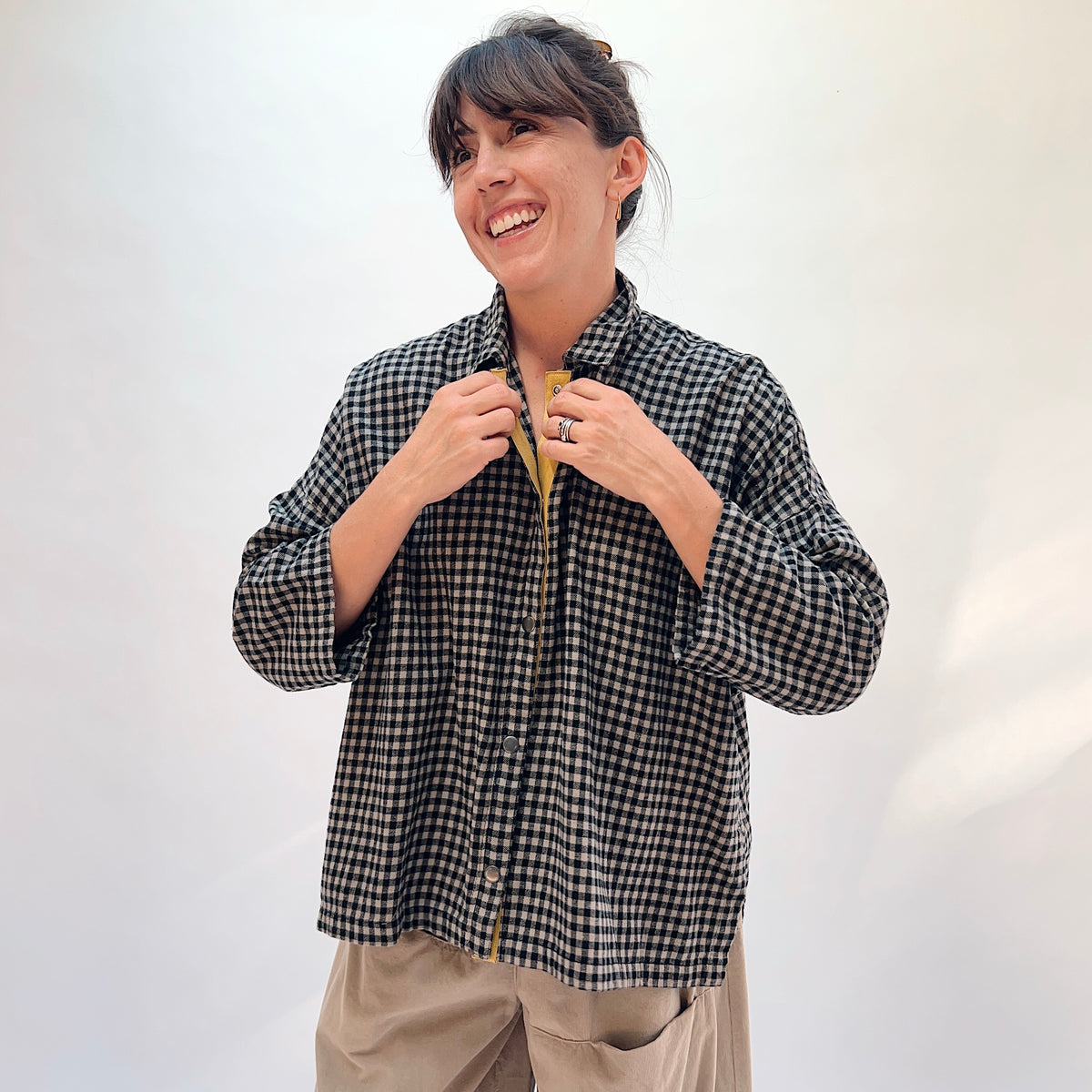 Model holding open the collar of the Eleven Stitch Small Check Big Shirt in Fennel, showing the mustard yellow contrast placket detail against the black and natural small gingham check cotton