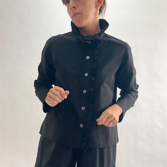 Full front view of model wearing Eleven Stitch black blossom collar shirt with hands on hips, showing ruffled blossom collar, ruffled button placket with shell buttons, long sleeves, relaxed boxy fit, and black color, styled with matching black wide-leg pants