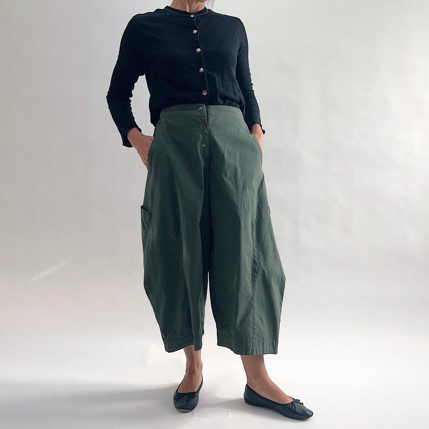 Full front view of model wearing Eleven Stitch konbu snap front pant with hands in pockets, showing snap front waistband, wide balloon leg, large patch pockets, gathered elastic hem, and deep forest green color, styled with black long sleeve button front cardigan and black ballet flats