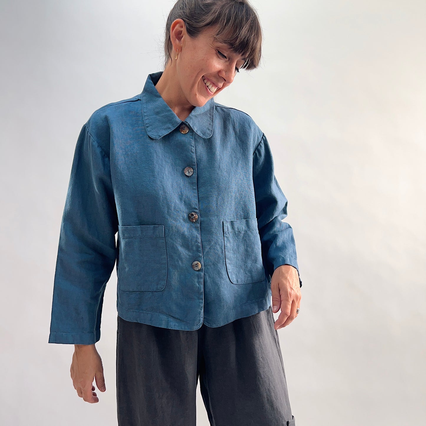 Model smiling and wearing the Bryn Walker Heavy Linen Audrey Jacket in Vitznau, front view showing patch pockets and shell button closure, paired with dark linen trousers