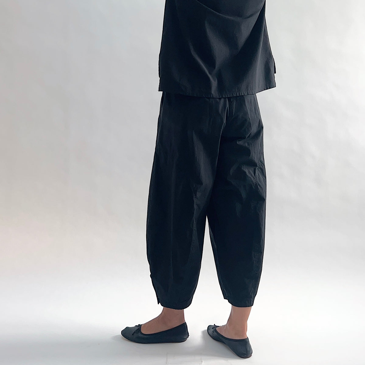Back view of model wearing Eleven Stitch black snap doreen pant, showing elastic waistband, wide balloon leg tapering to snap hem at ankle, side seam detailing, and black color, styled with matching black top and black ballet flats