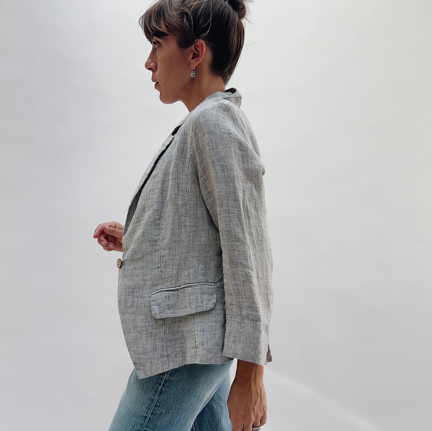 Second side profile view of the Linen Blazer in Grey Pinstripe by Yuvita, showing the open-front drape, structured shoulder, and flap pocket detail in grey pinstripe linen, styled with blue jeans