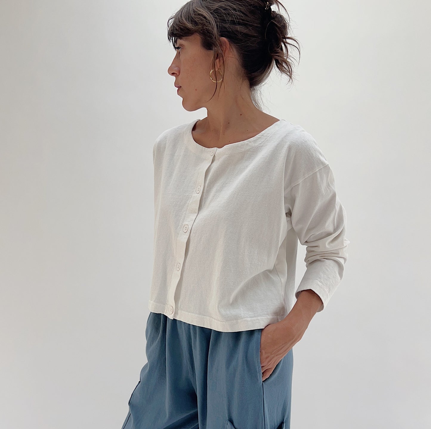 Side three-quarter view of model wearing Pacific Cotton cream button-front shirt, showing crew neckline, button placket, long sleeves, and cropped length layered over blue pants