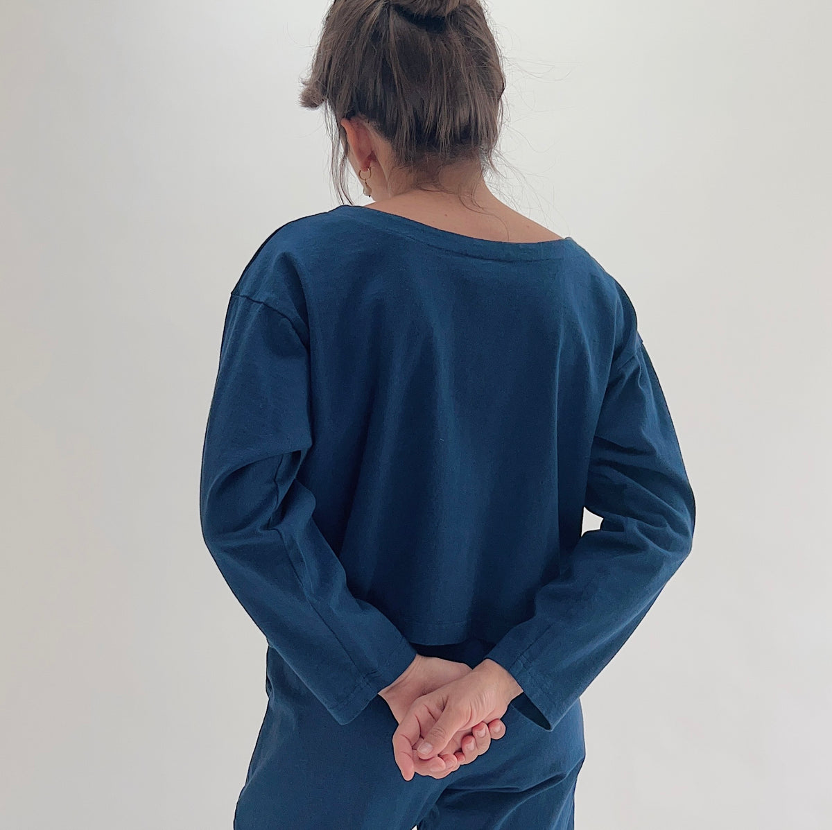 Back view of model wearing Pacific Cotton nova long sleeve button front shirt, showing wide back neckline, long sleeves, boxy cropped silhouette, banded hem, and deep teal color