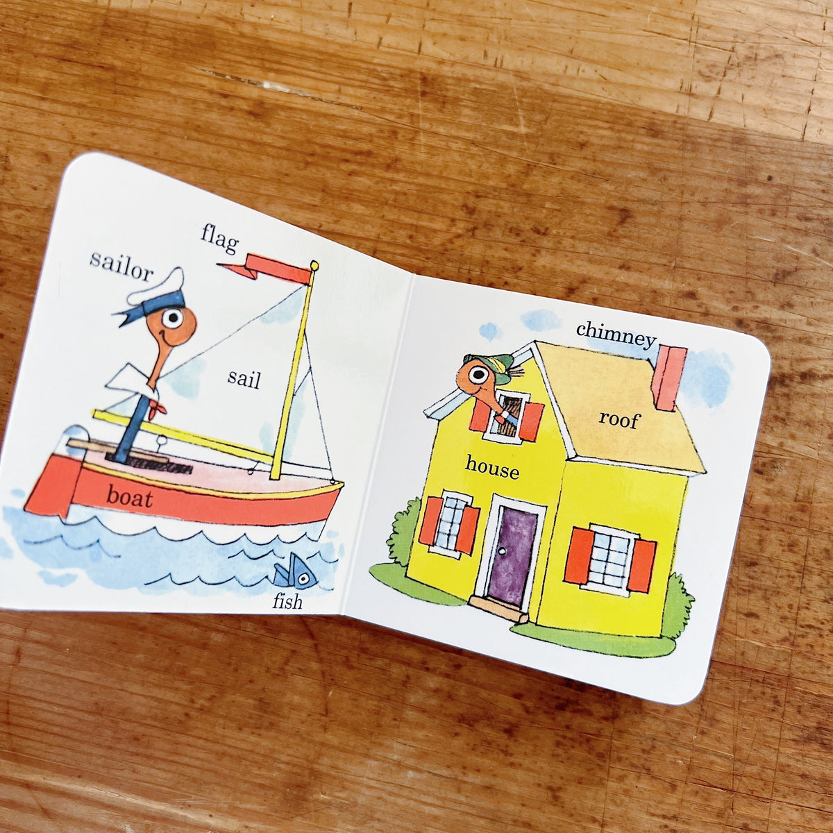 Interior spread of Richard Scarry's Lowly Worm Word Book showing labeled illustrations: Lowly Worm as a sailor on a red boat with words flag, sail, boat, and fish on the left page, and Lowly Worm peeking from a yellow house labeled with chimney, roof, and house on the right page