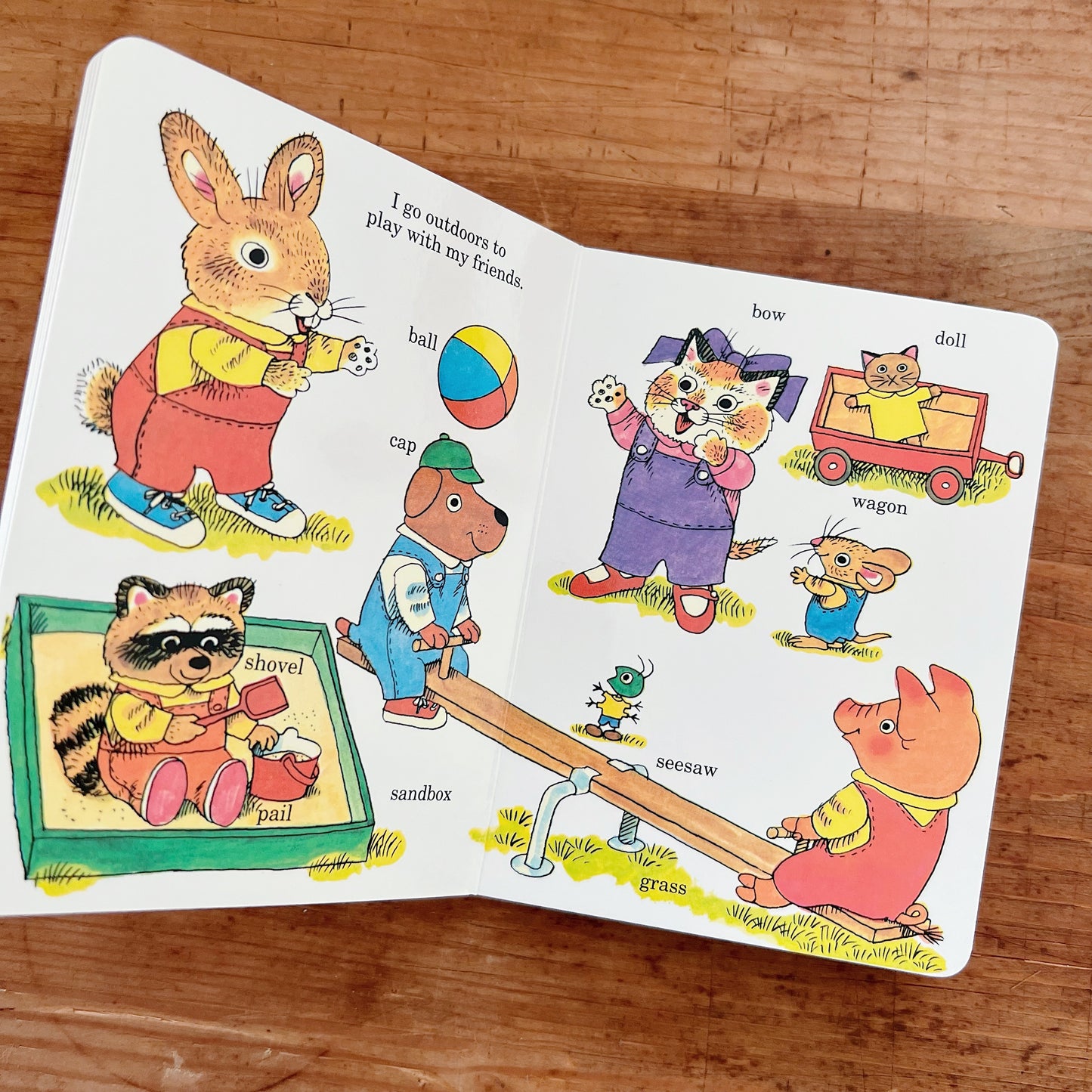 Open interior spread of Richard Scarry's Best Little Board Book Ever showing the playground page from the Early Words section, with white background and Richard Scarry's color illustrations of animal characters at play. Header text reads "I go outdoors to play with my friends." Labeled objects include: Frannie the bunny (upper left), a raccoon in a sandbox with a shovel and pail, a dog in a cap, a colorful ball, a cat with a bow in a purple dress waving, a small mouse in blue overalls, a pig on a seesaw, a