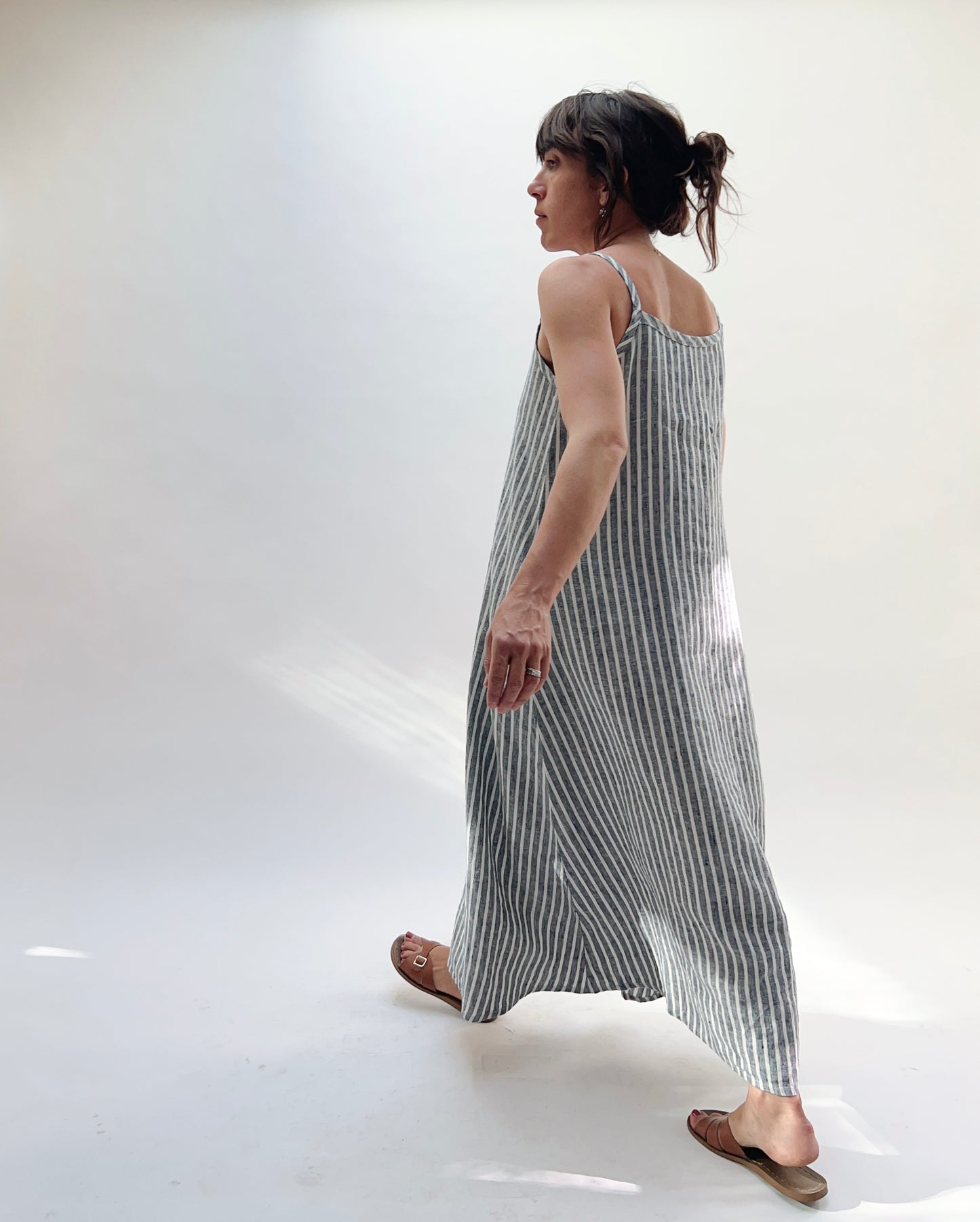 Woman wearing a long, striped linen dress in white and grey on a white background as seen from behind.